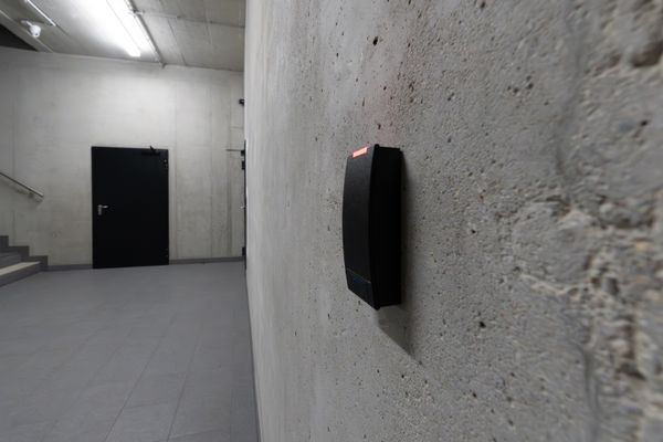 A wall-mounted access reader on bare concrete, its red status light lit, with a black door and a stairwell down the corridor behind