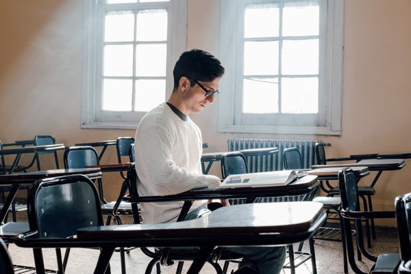 A man in a white sweater and glasses reading at a school desk in an empty classroom, rows of empty desks around him