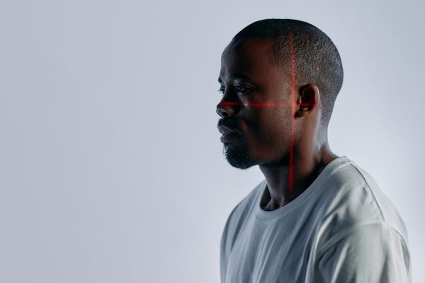 A man in profile against a pale grey studio background, a thin red laser crosshair laid across his cheekbone and temple